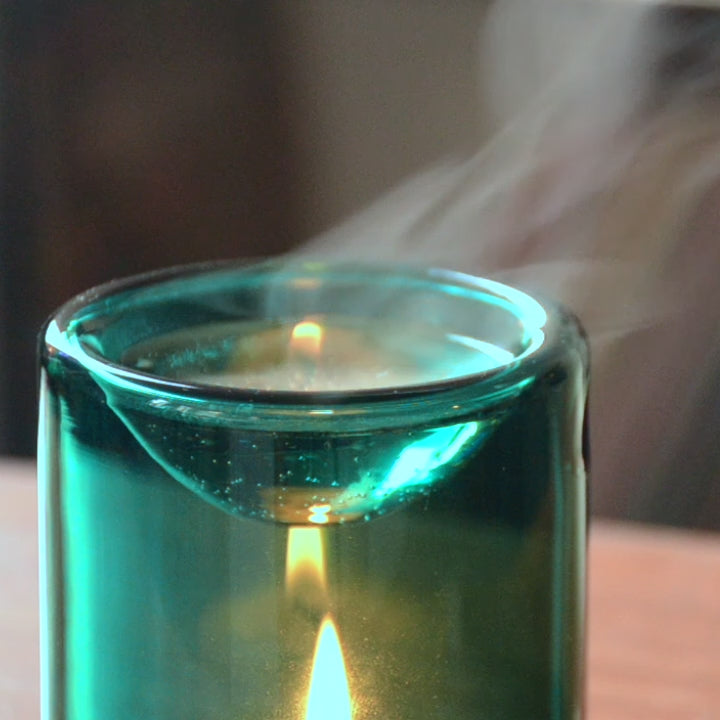 Marga glass aroma diffuser in turquoise releasing soft vapor with a gentle candle flame in a calm setting